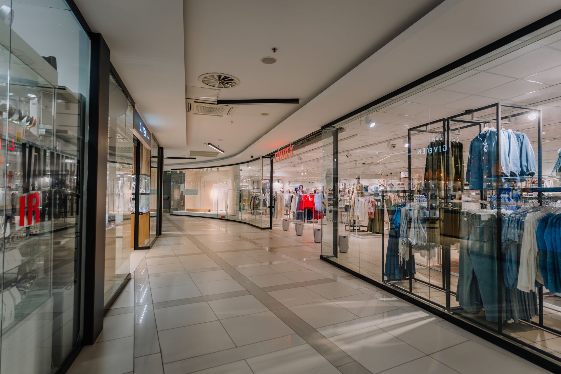 A brightly lit, wide indoor shopping mall corridor with glass storefronts displaying clothing on racks and shelves.