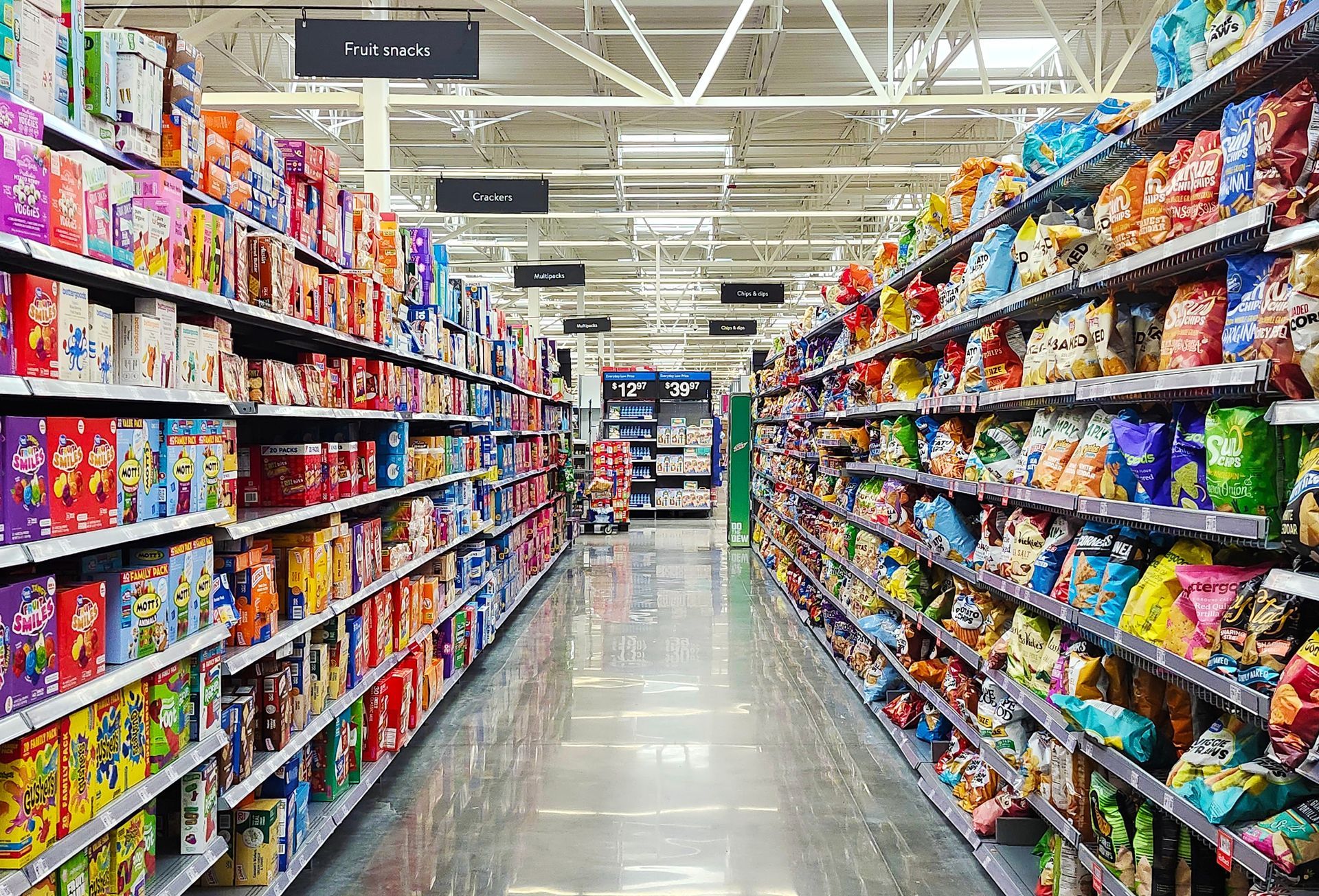 A grocery store aisle with tall shelves stocked with various colorful boxes and snack bags, stretching into the distance.