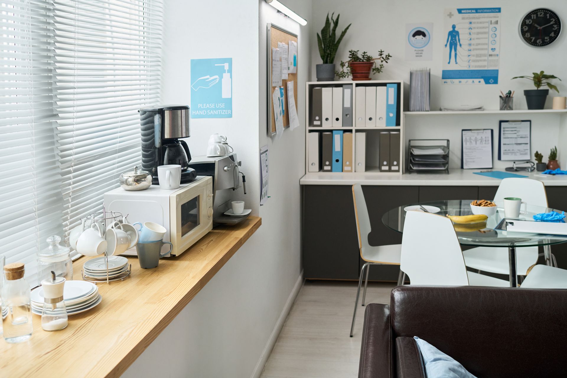 A coffee station on a wooden surface with a tray of snacks, containers, a small potted plant, and stacks of paper cups.
