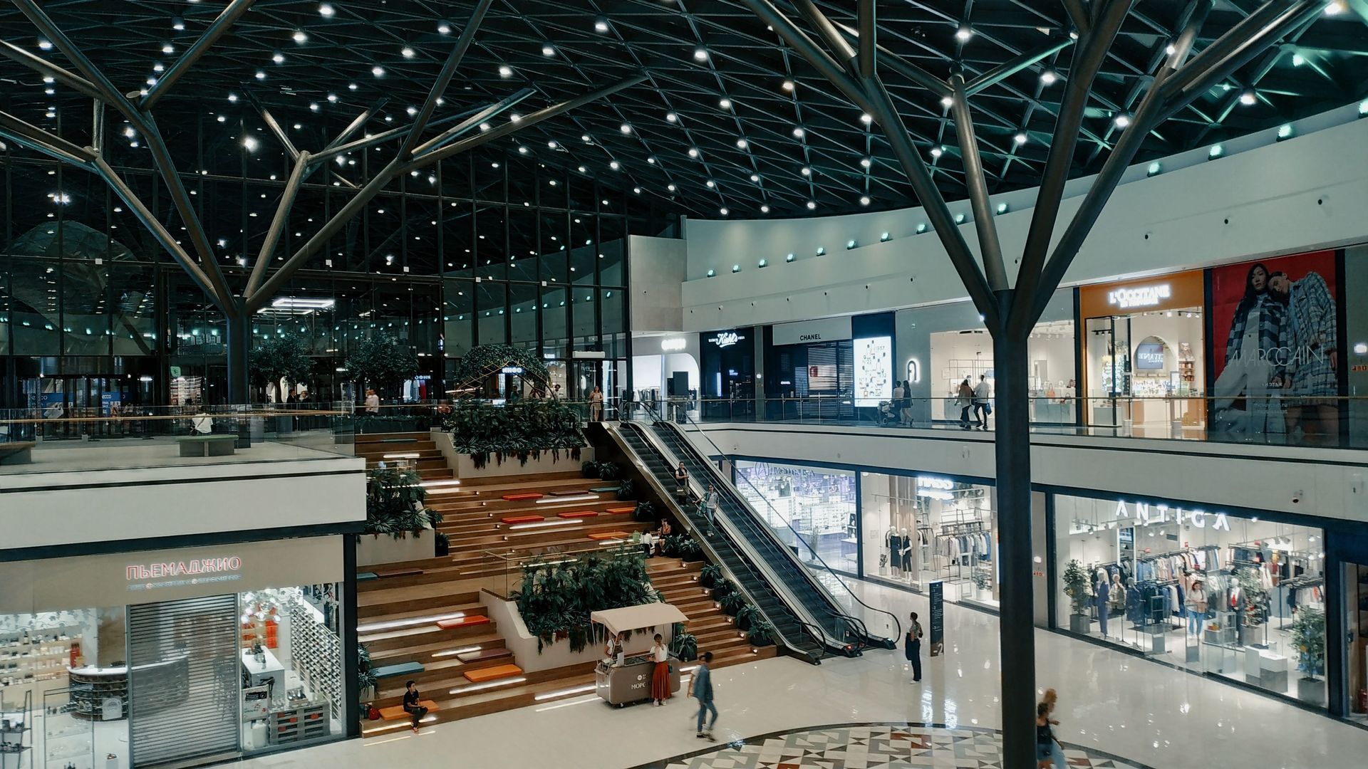 Modern shopping mall interior with escalators, glass storefronts, and a few people walking beneath a high ceiling