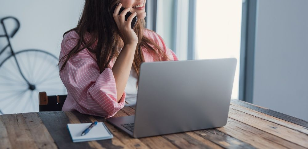 Woman speaking on a phone beside a laptop at a wooden desk, with a bicycle in the background.