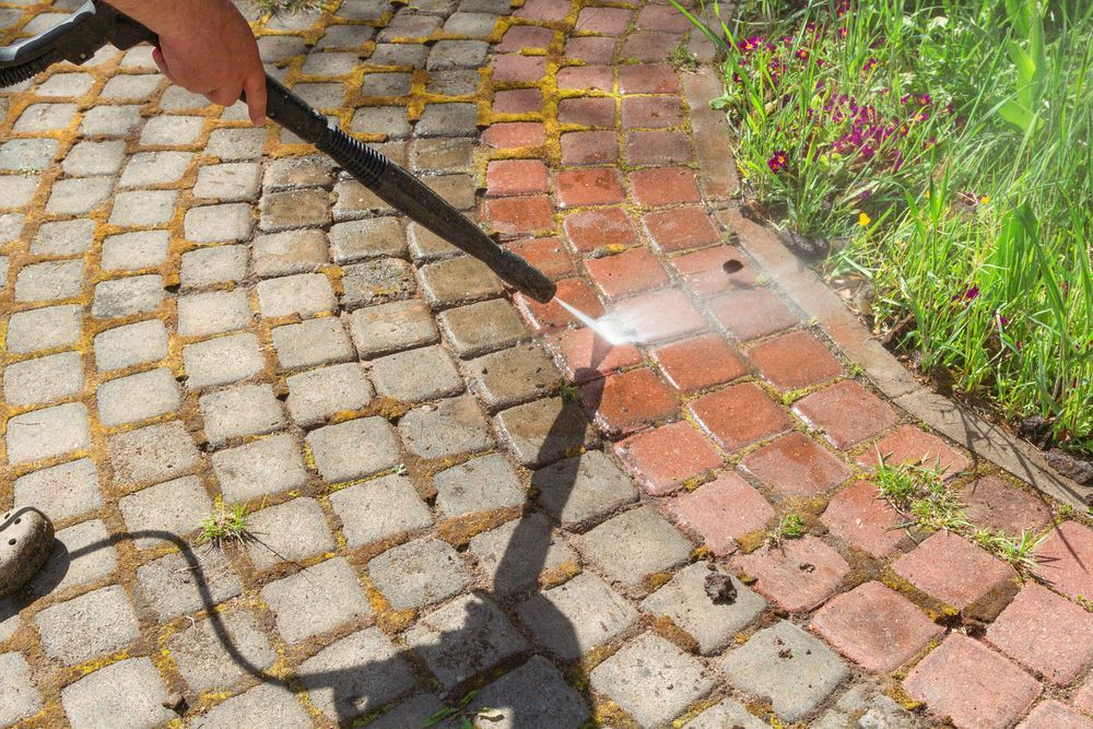 A power washer sprays water onto a metal ventilation grate set into a concrete wall.