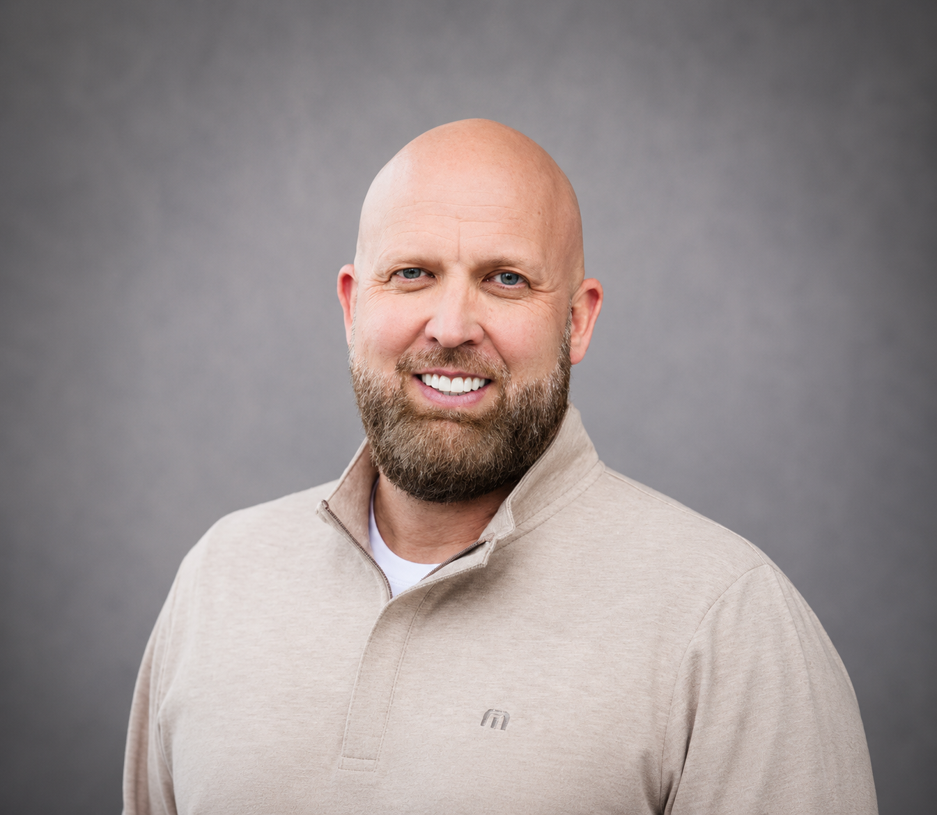 Smiling bald man with a beard wearing a beige sweater against a gray background