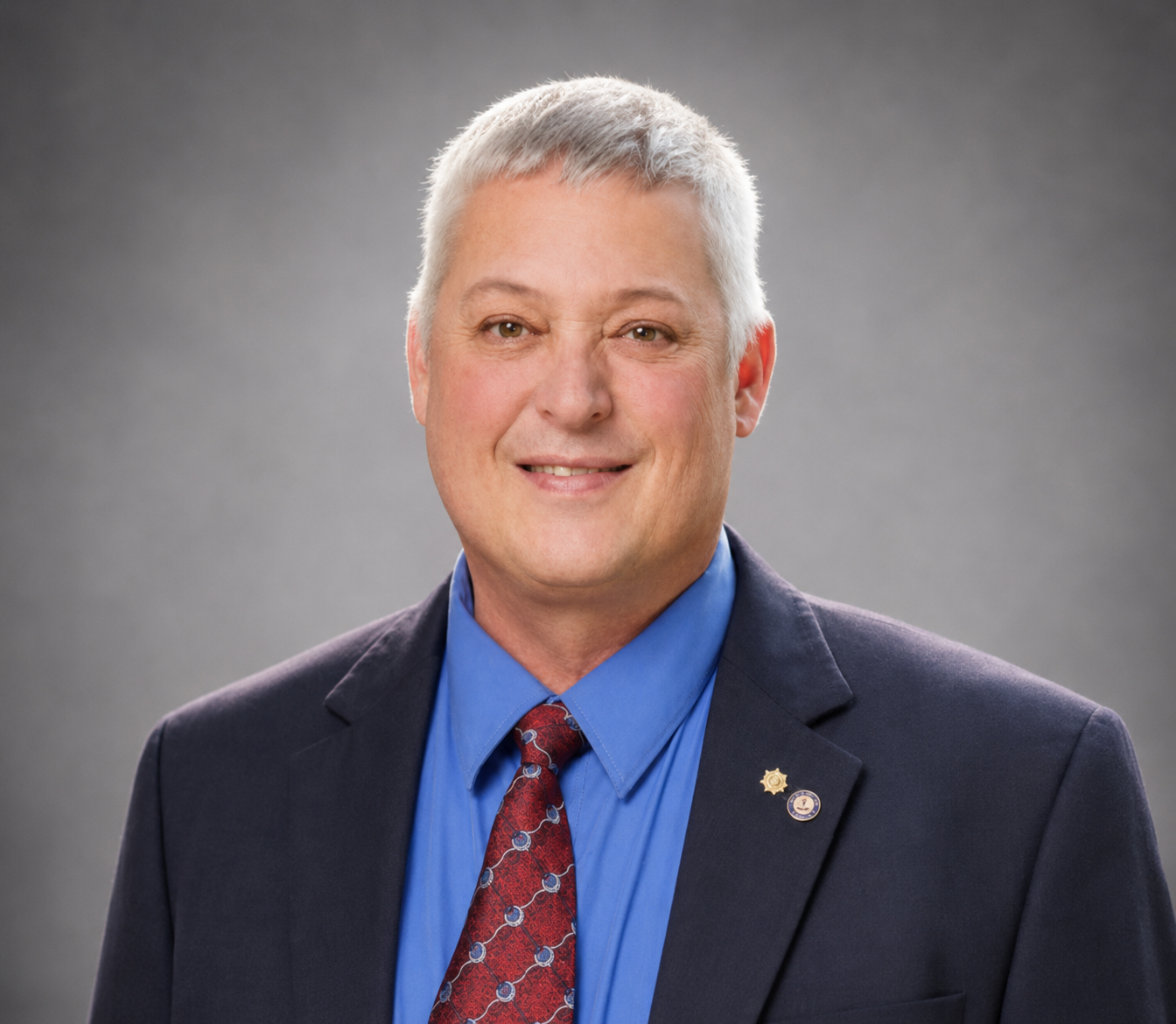 Portrait of a smiling person in a navy suit, blue shirt, and red tie against a gray backdrop
