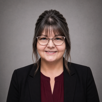 Smiling woman with glasses and wavy brown hair in a black blazer and burgundy top against a gray background