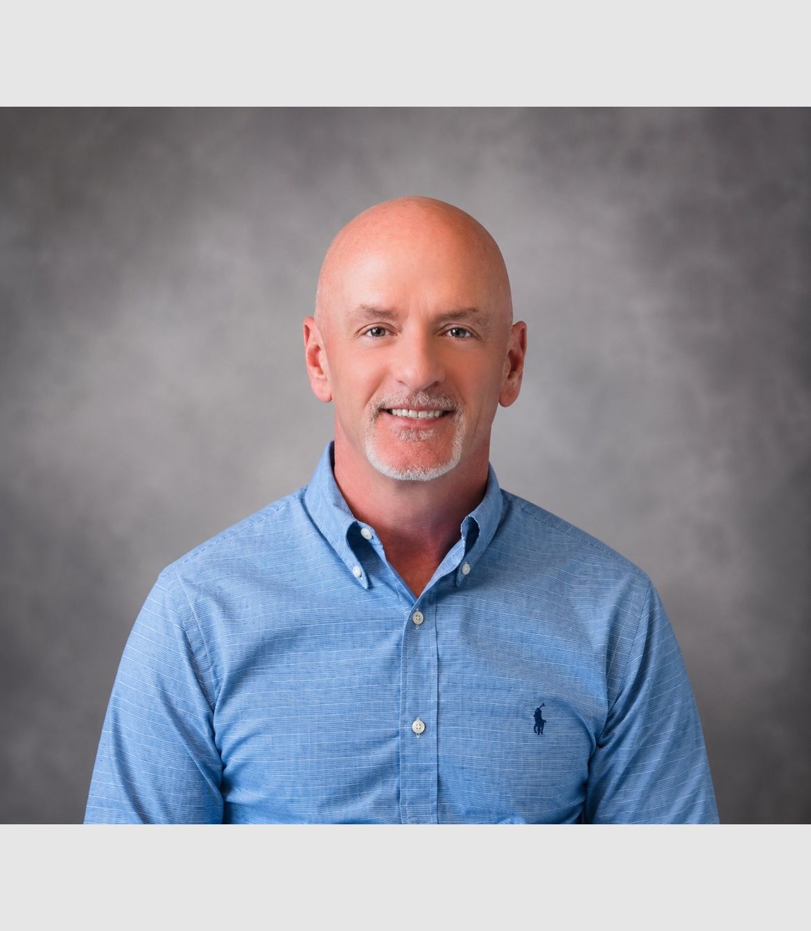 Portrait of a smiling bald man in a blue button-down shirt against a gray studio backdrop