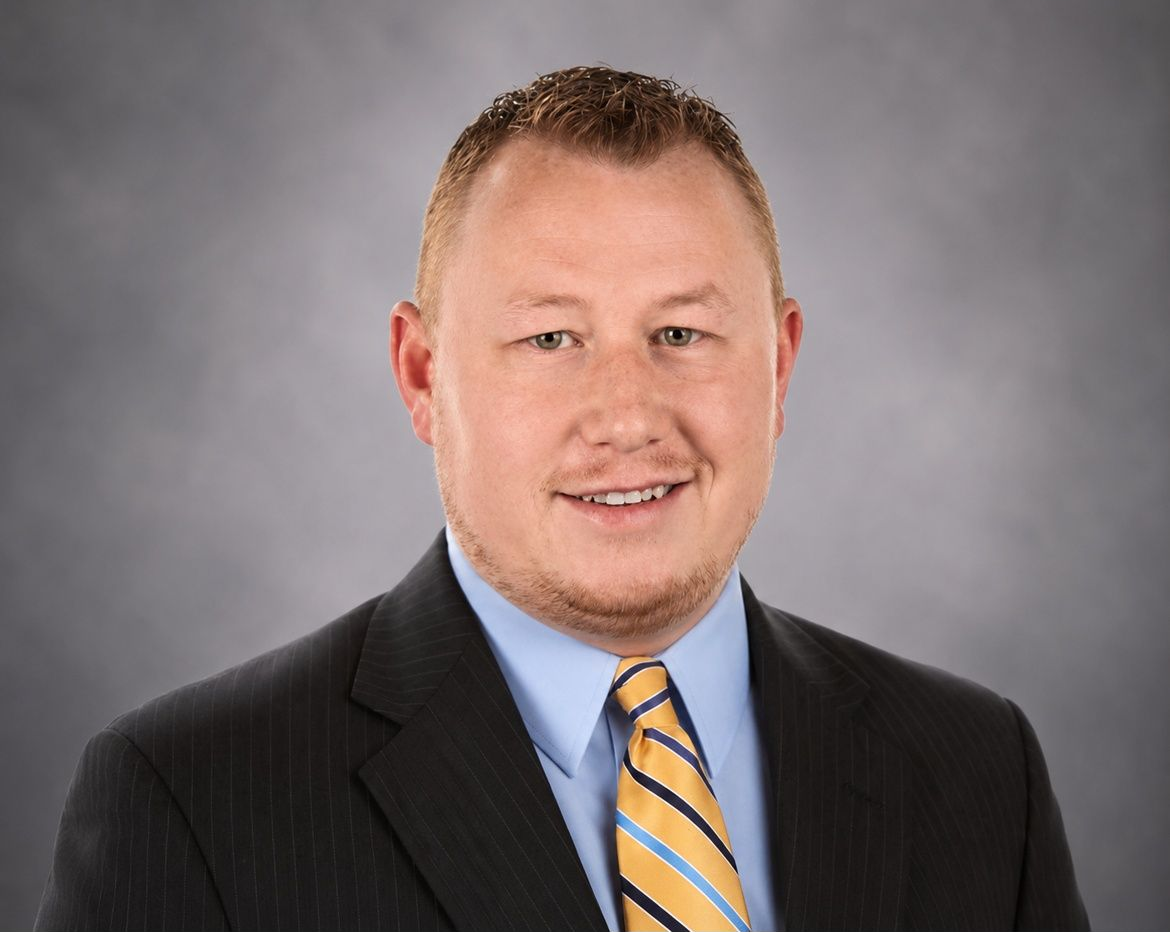 Formal headshot of a smiling person in a dark suit, blue shirt, and yellow striped tie against a gray background