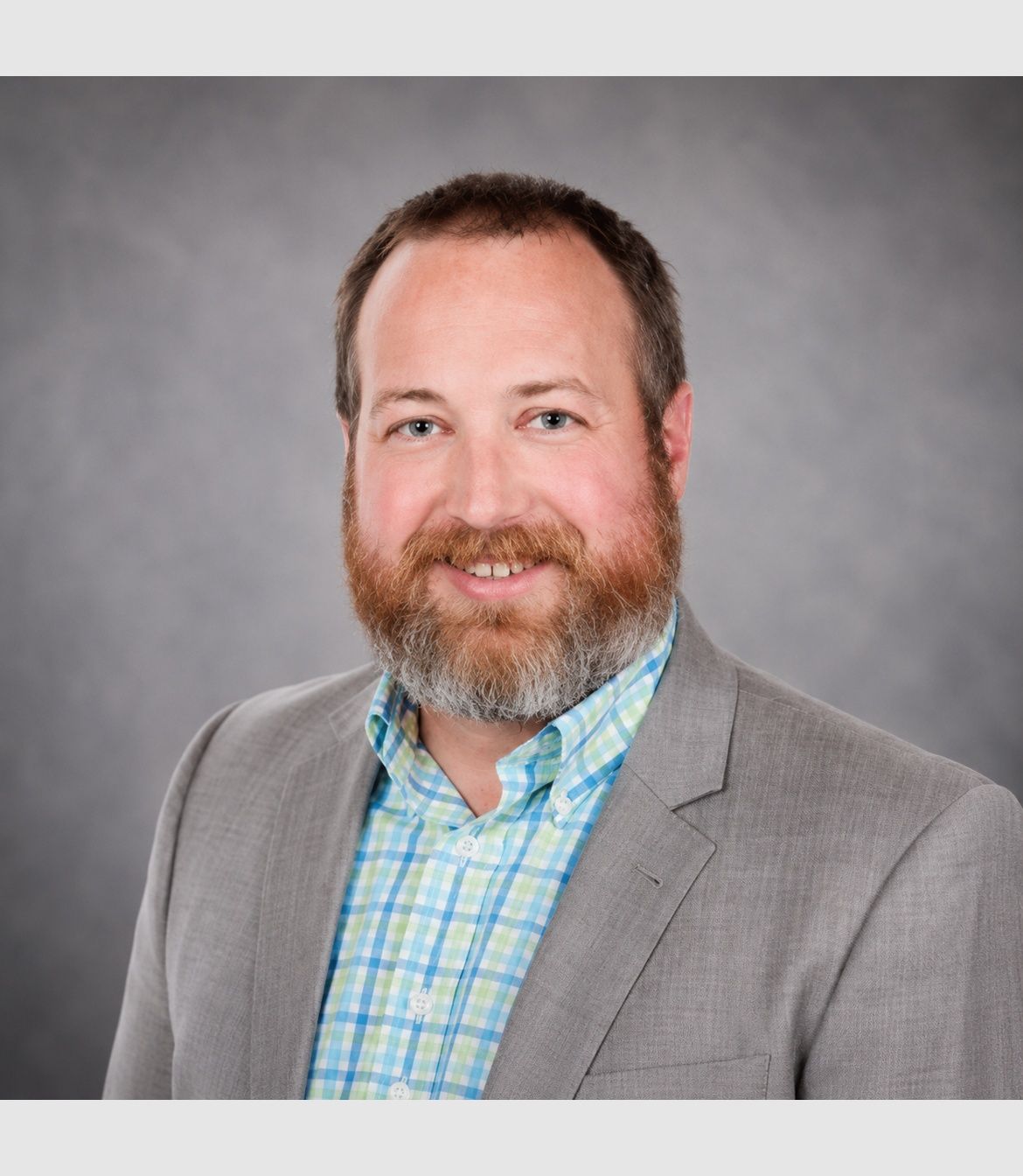 Portrait of a smiling man in a gray blazer and blue plaid shirt against a gray backdrop