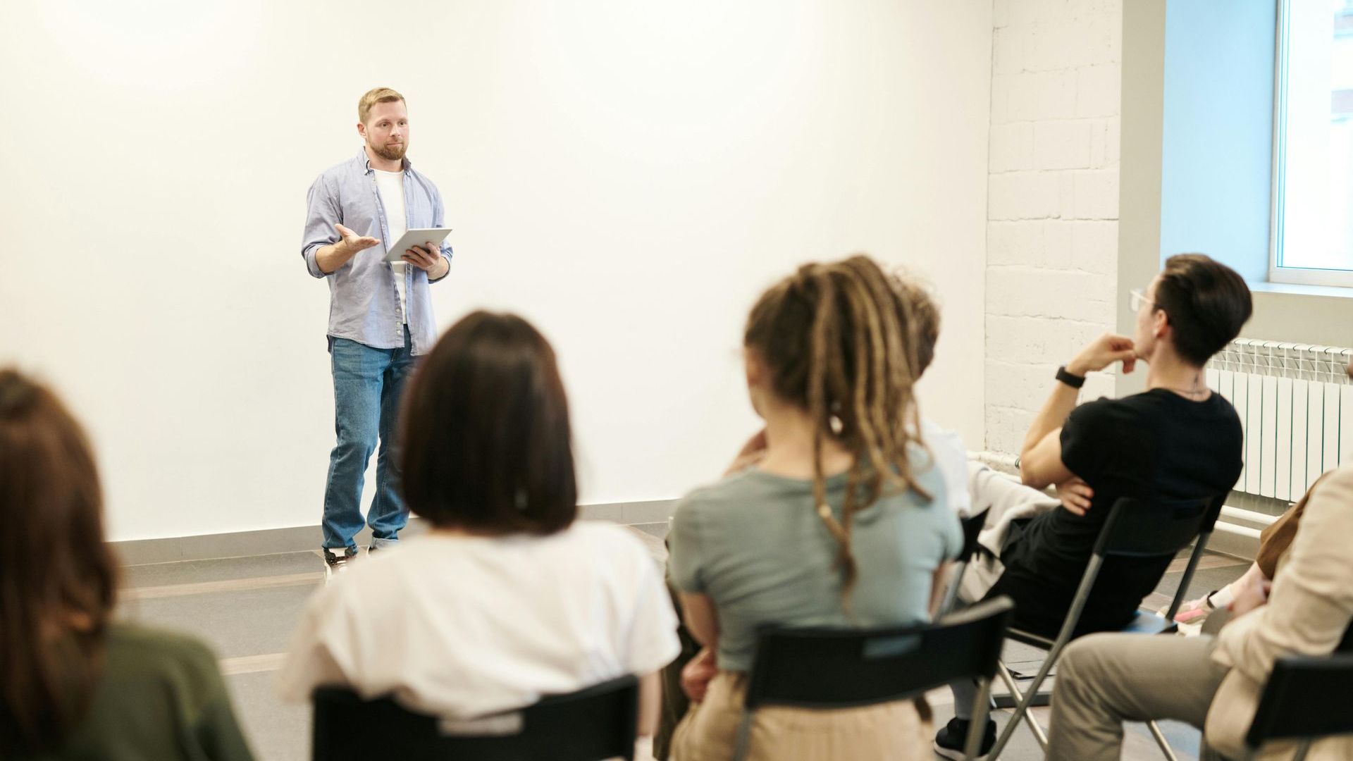 Speaker presenting to seated audience in a bright room, with a projected screen beside him.