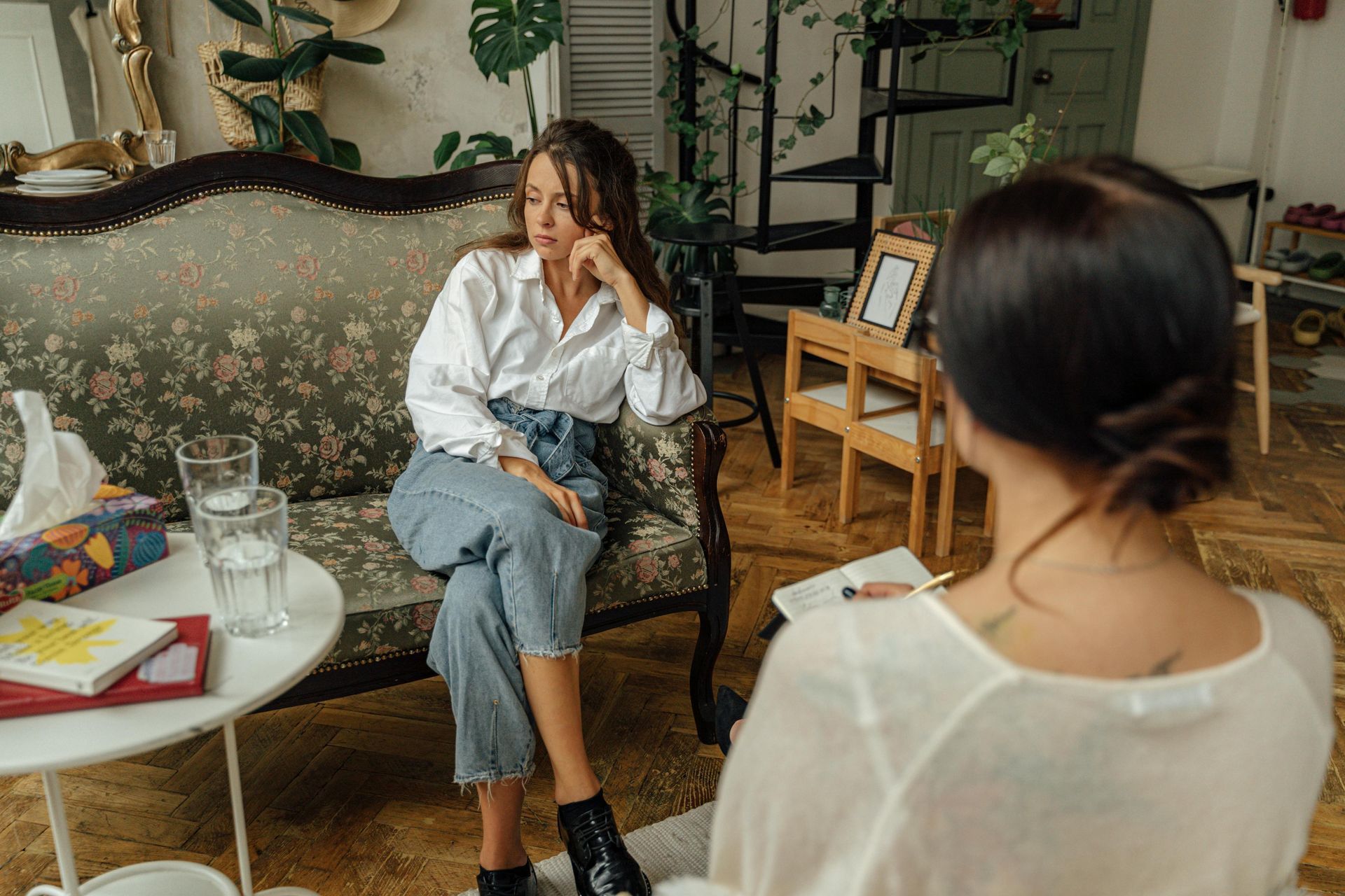Two people talking in a cozy living room with vintage furniture and a small table with drinks and notebooks