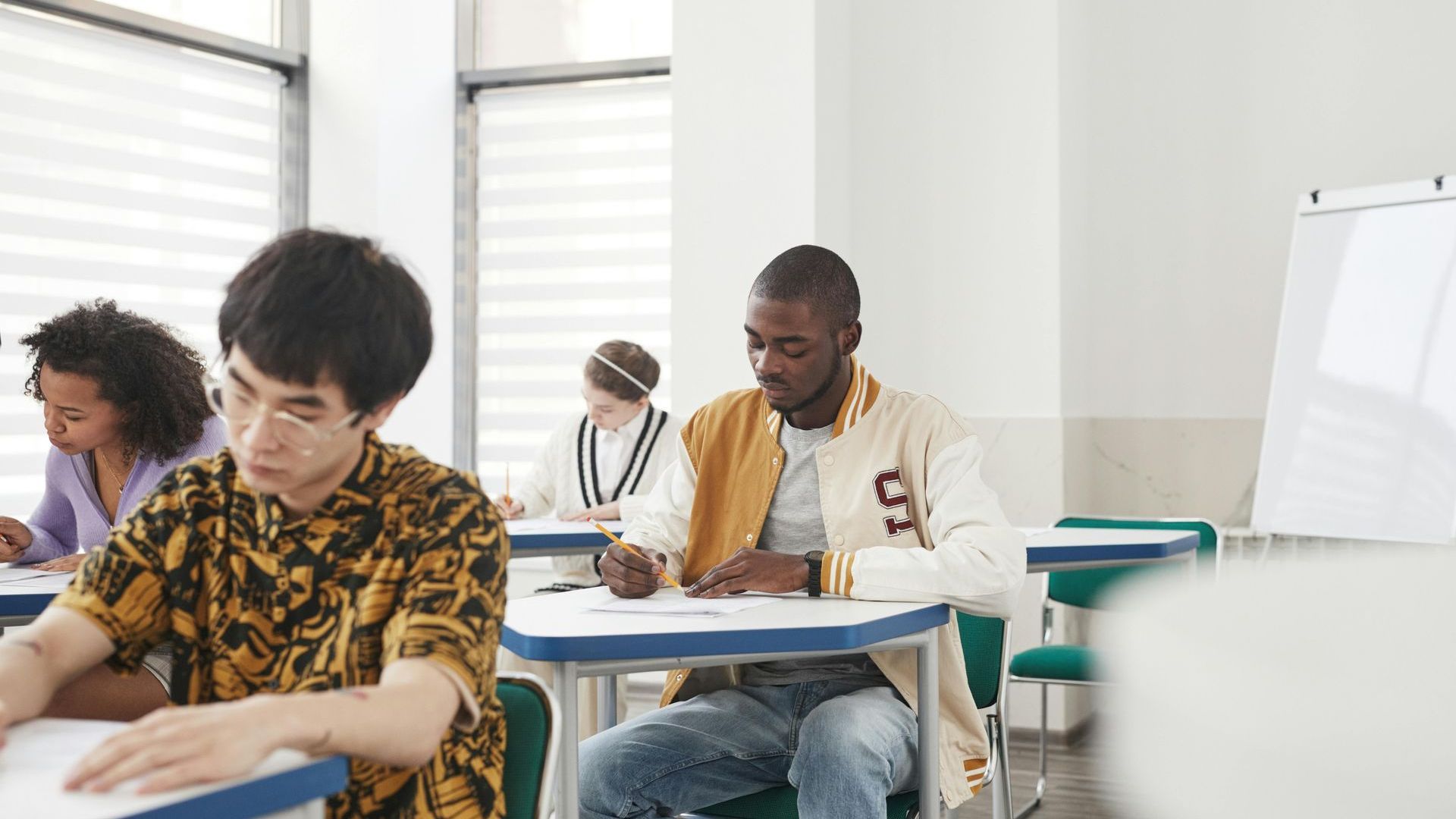 Students writing at desks in a bright classroom with a whiteboard and window blinds.