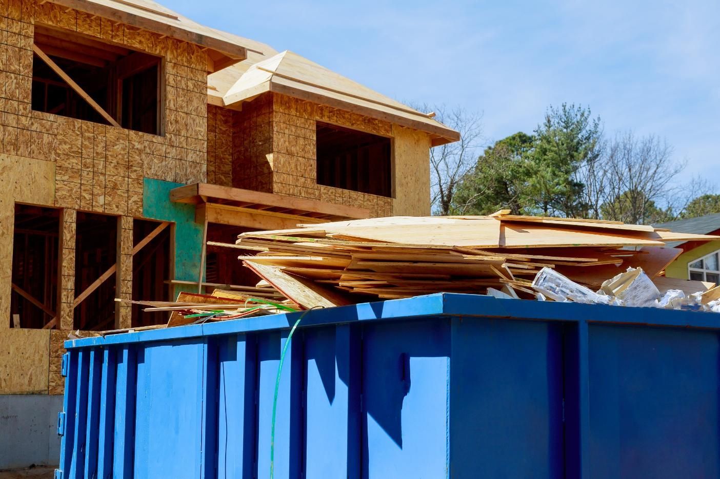 Blue dumpster in front of a wooden house under construction with lumber stacked on top
