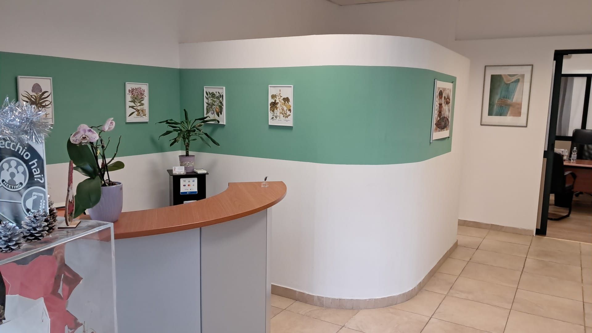 Area reception con parete curva bianca e verde, scrivania e stampe d'arte botanica.