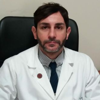 Medico in camice bianco, seduto alla scrivania, che guarda la telecamera. Capelli scuri, barba, cravatta.