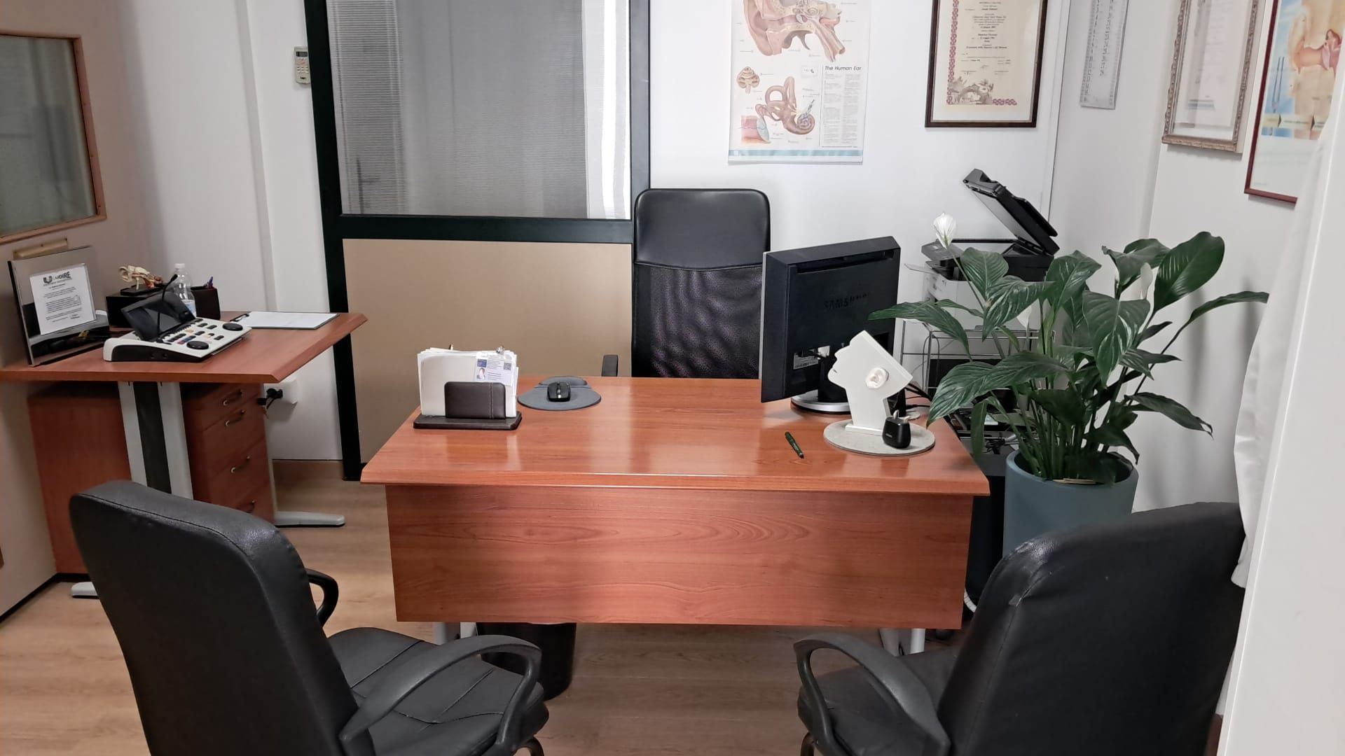 Ufficio con scrivania, sedie, computer e cartelle cliniche alla parete.