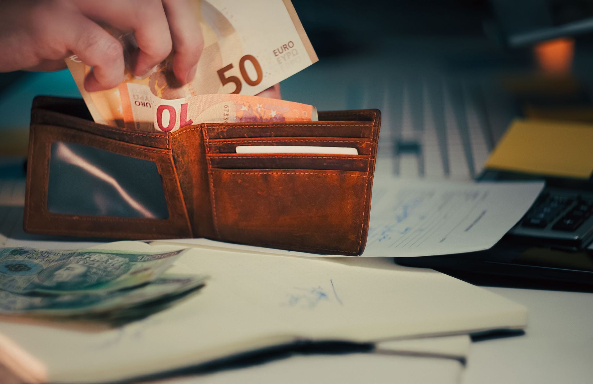Hand placing euro banknotes into an open brown wallet on a desk with papers and a calculator
