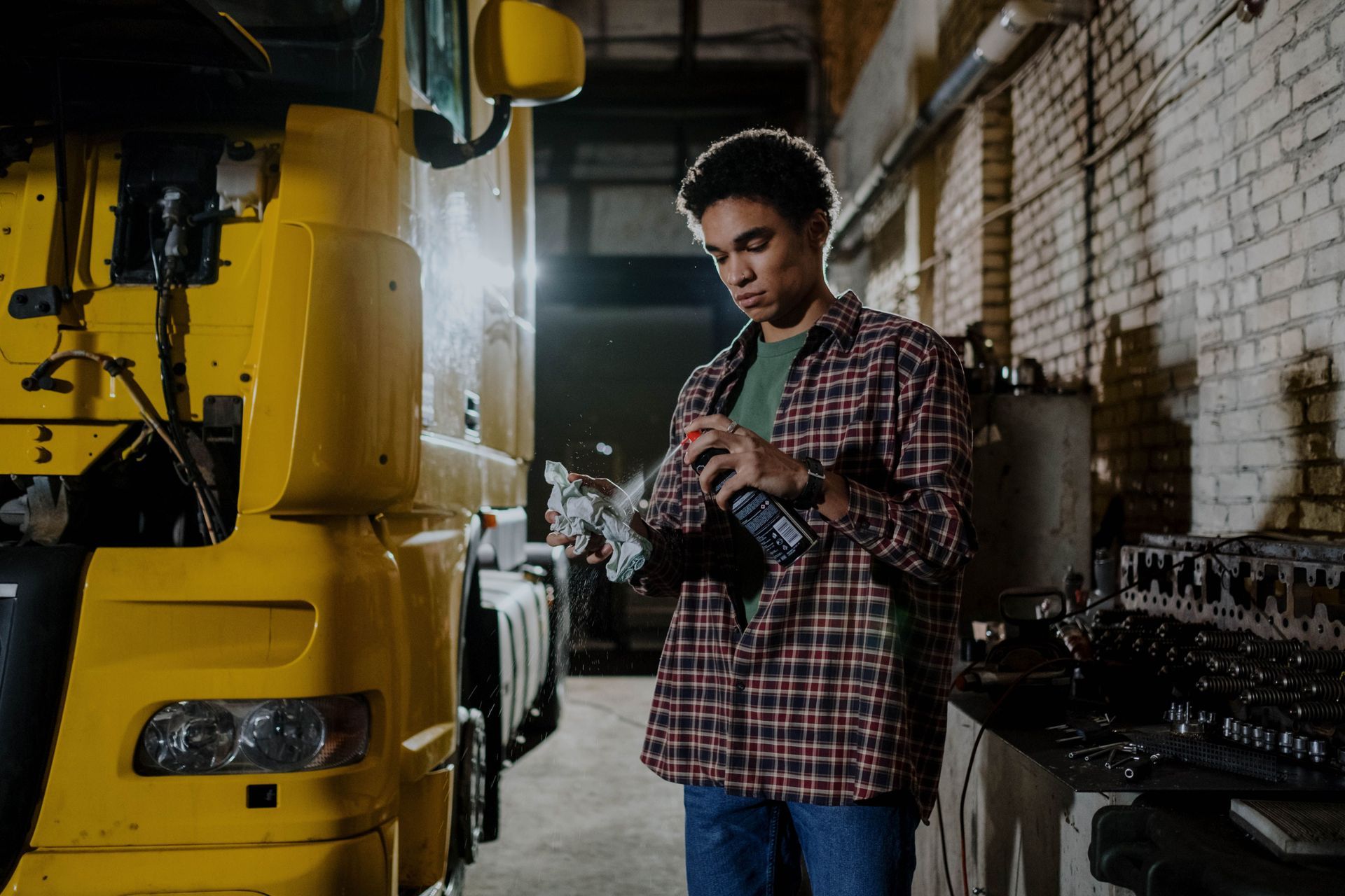 Mechanic inspecting a car part beside a yellow truck in a dim garage