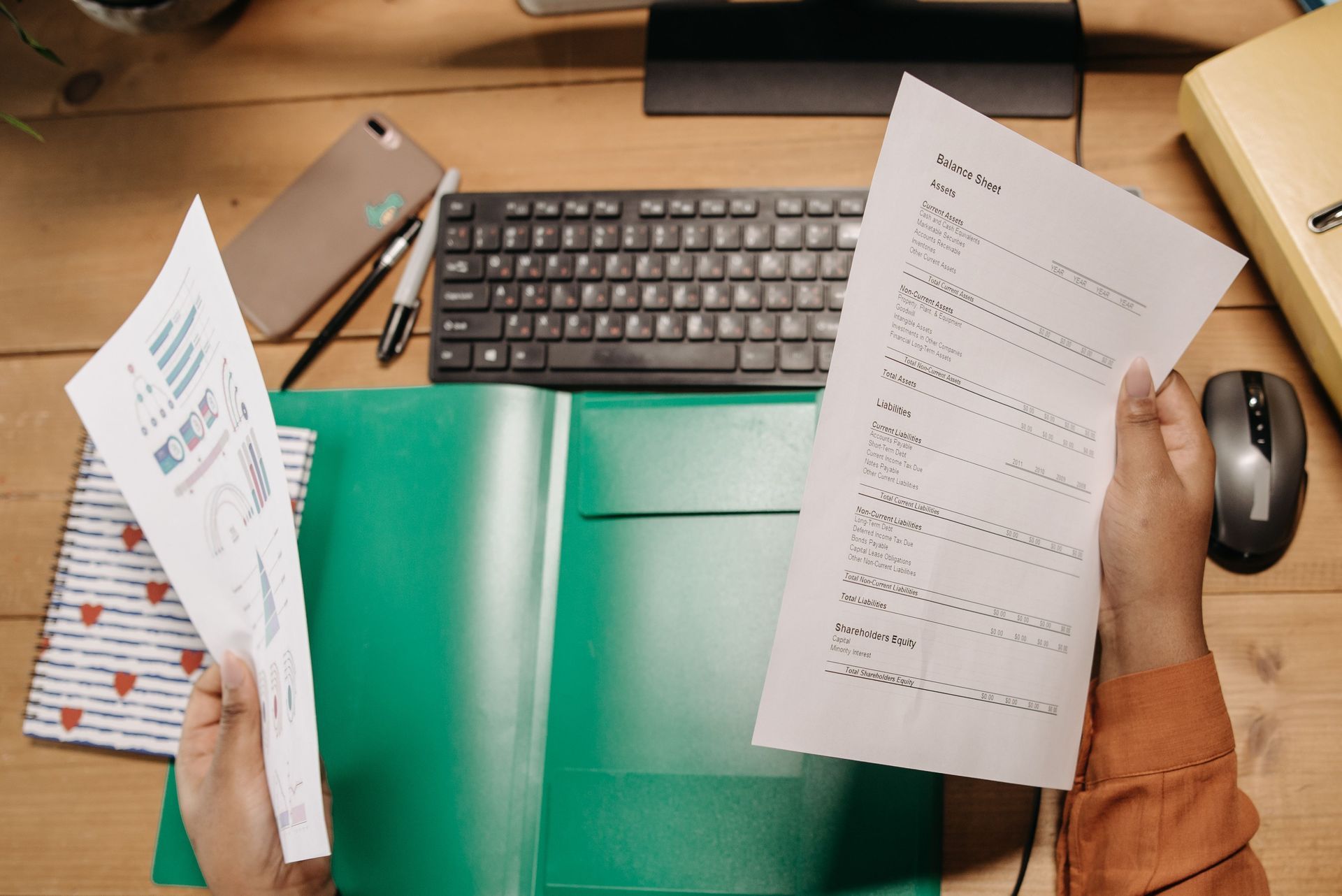 Desk with keyboard, laptop, paperwork, green folder, and hands holding a document.
