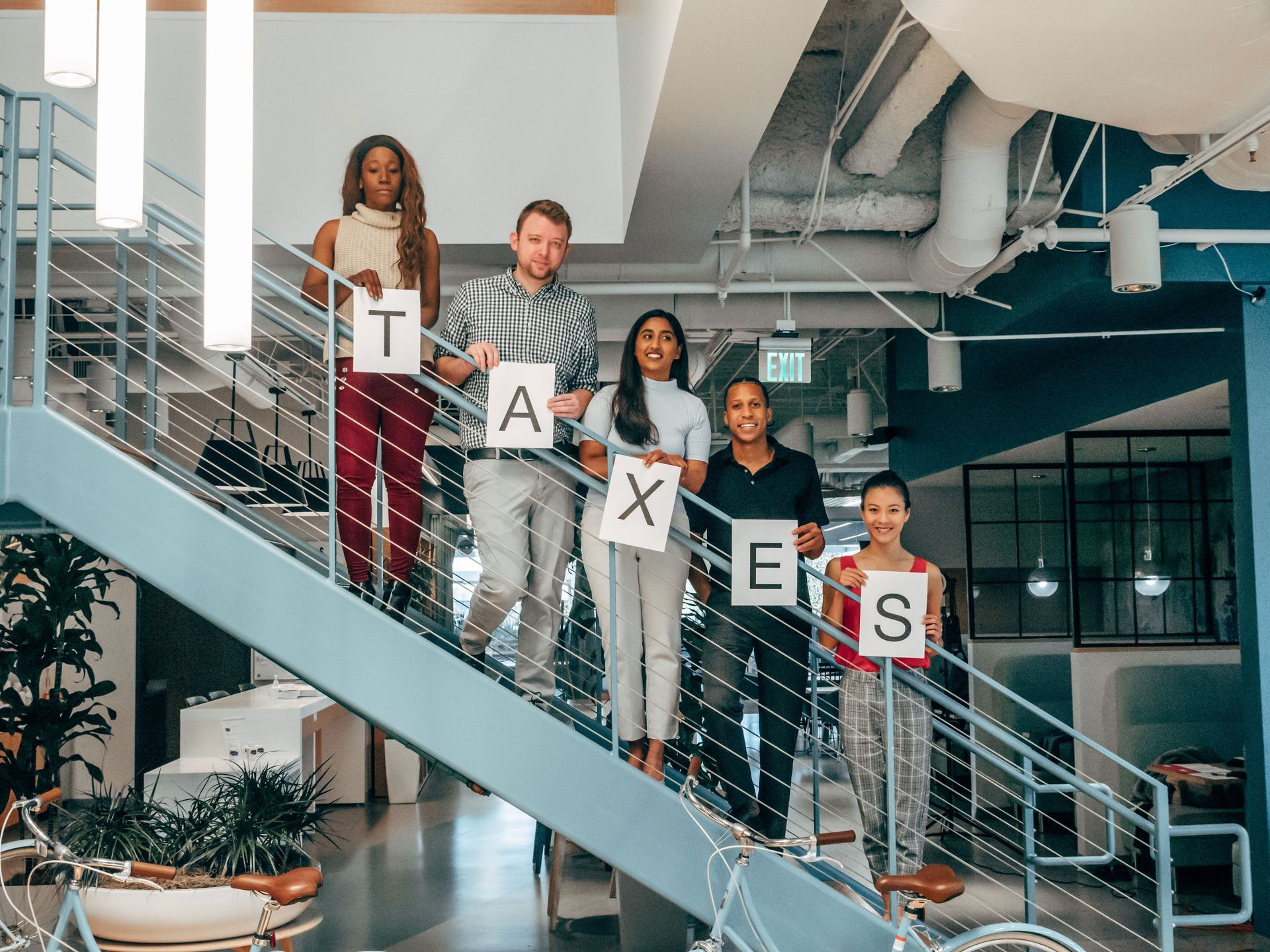 Five people on a stairway hold signs spelling “TAXES” in an office atrium.
