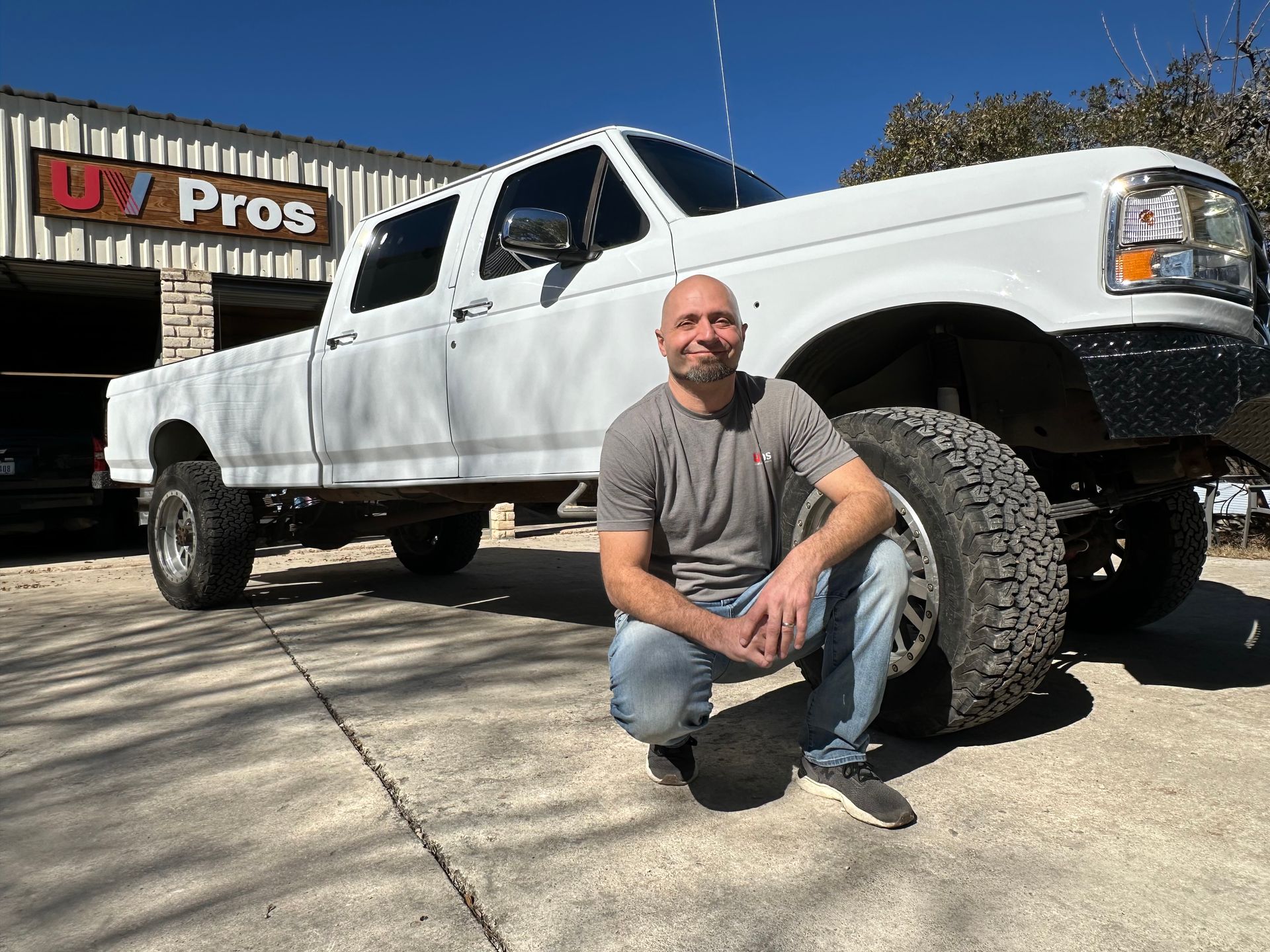 Man kneels in front of a white lifted truck with UT Pros building in the background.