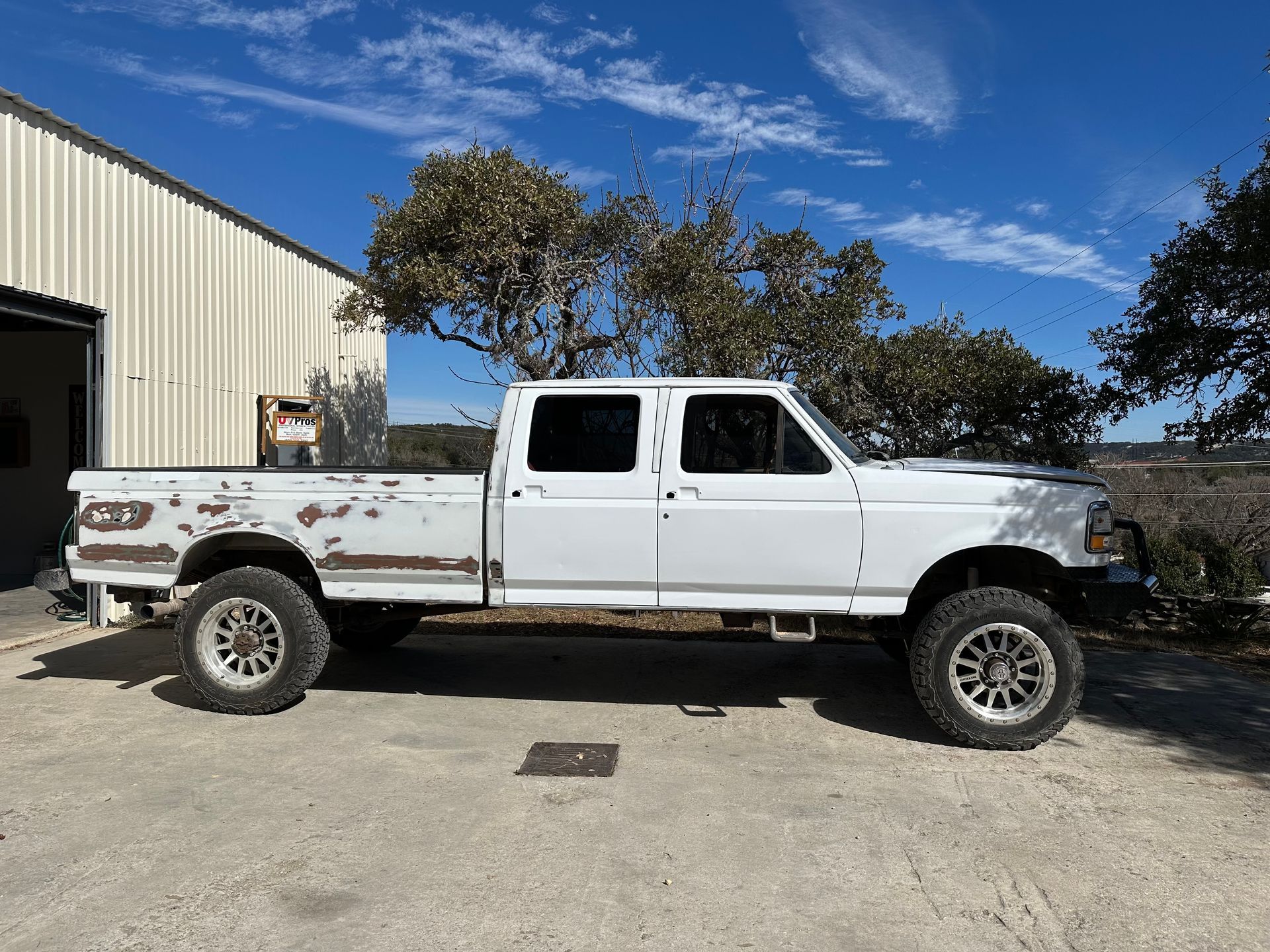 White, lifted, extended-cab pickup truck parked outside a metal building on a sunny day.