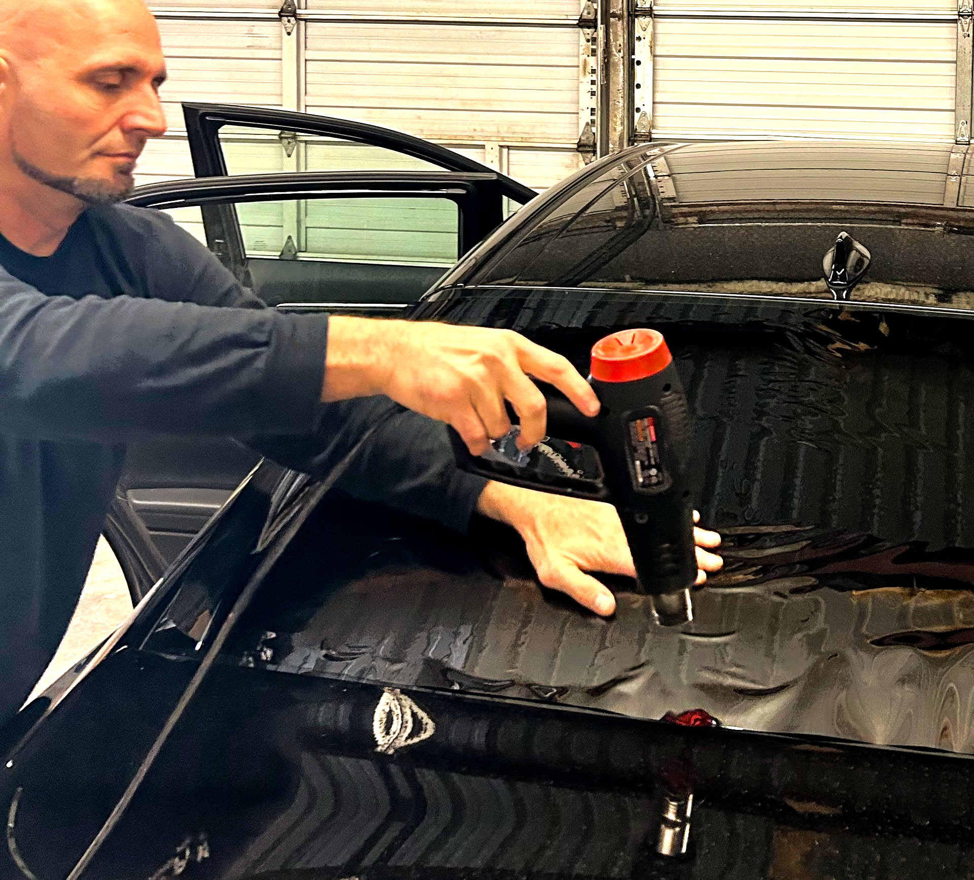 Man using a heat gun to apply film to a black car's window in a garage.