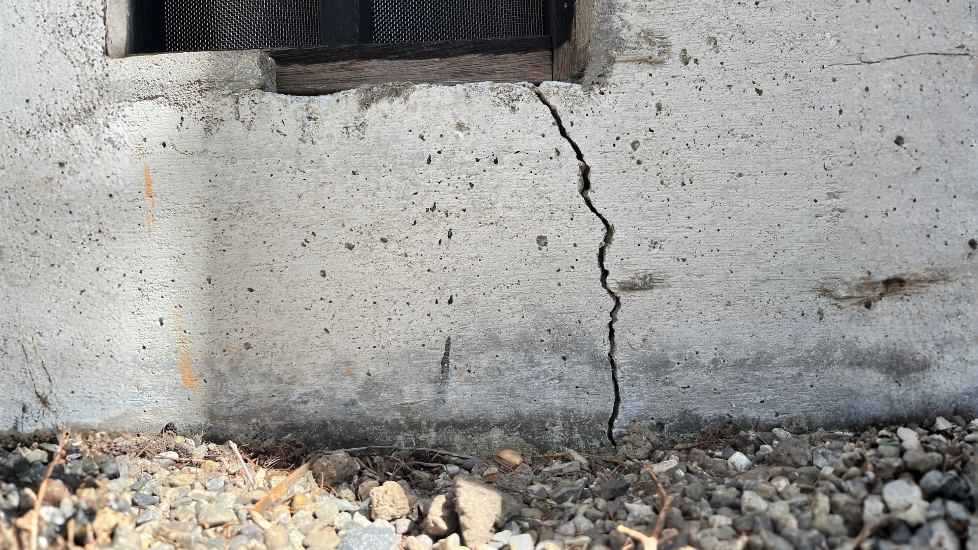 Cracked concrete wall beneath a vent, with gravel at the base.