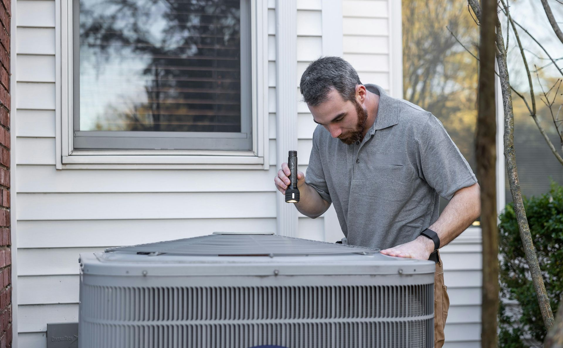 Man inspecting an air conditioning unit outside a house, holding a flashlight.