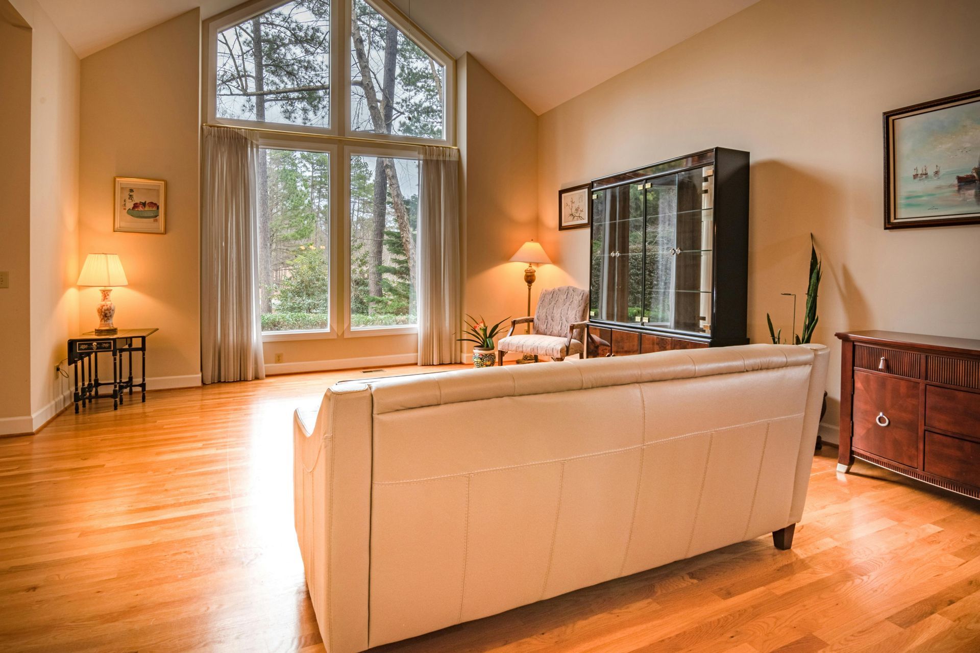Spacious living room with hardwood floors, large window, leather couch, cabinet, and lamps.