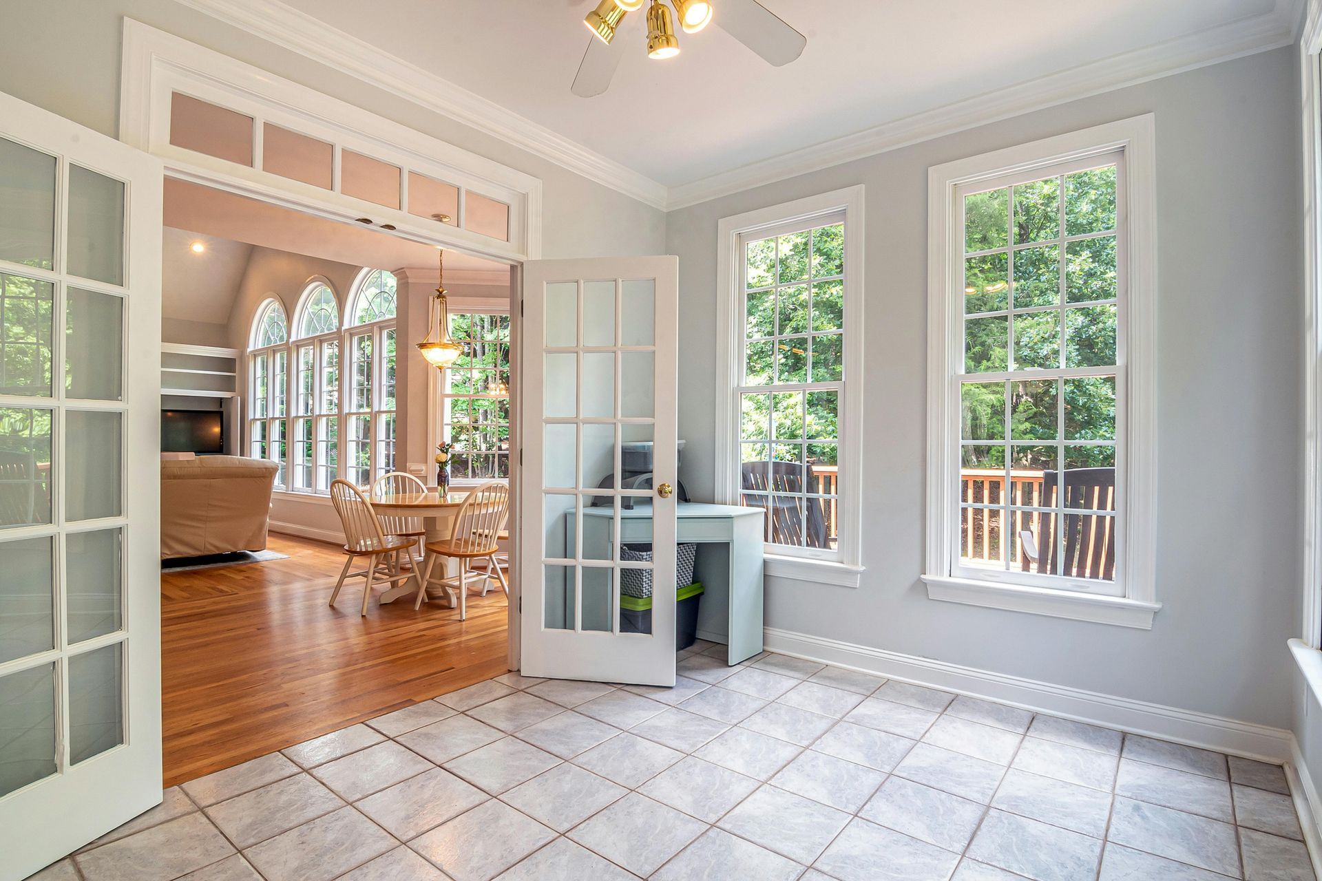 Sunroom with tile floor, glass doors, and windows overlooking greenery; connects to a living area with hardwood floors.