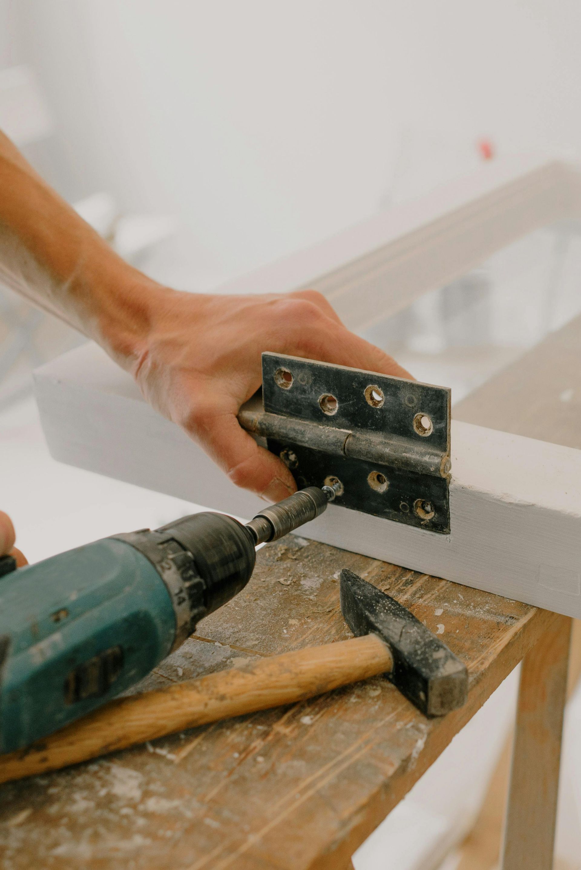 Hand using a power drill to attach a hinge to a white wooden frame; a hammer rests nearby.