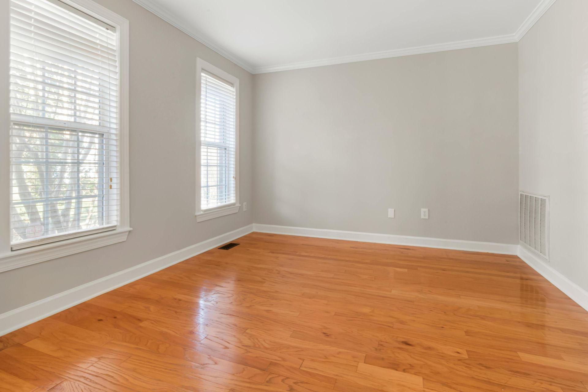 Empty room with hardwood floors, two windows with blinds, and light gray walls.