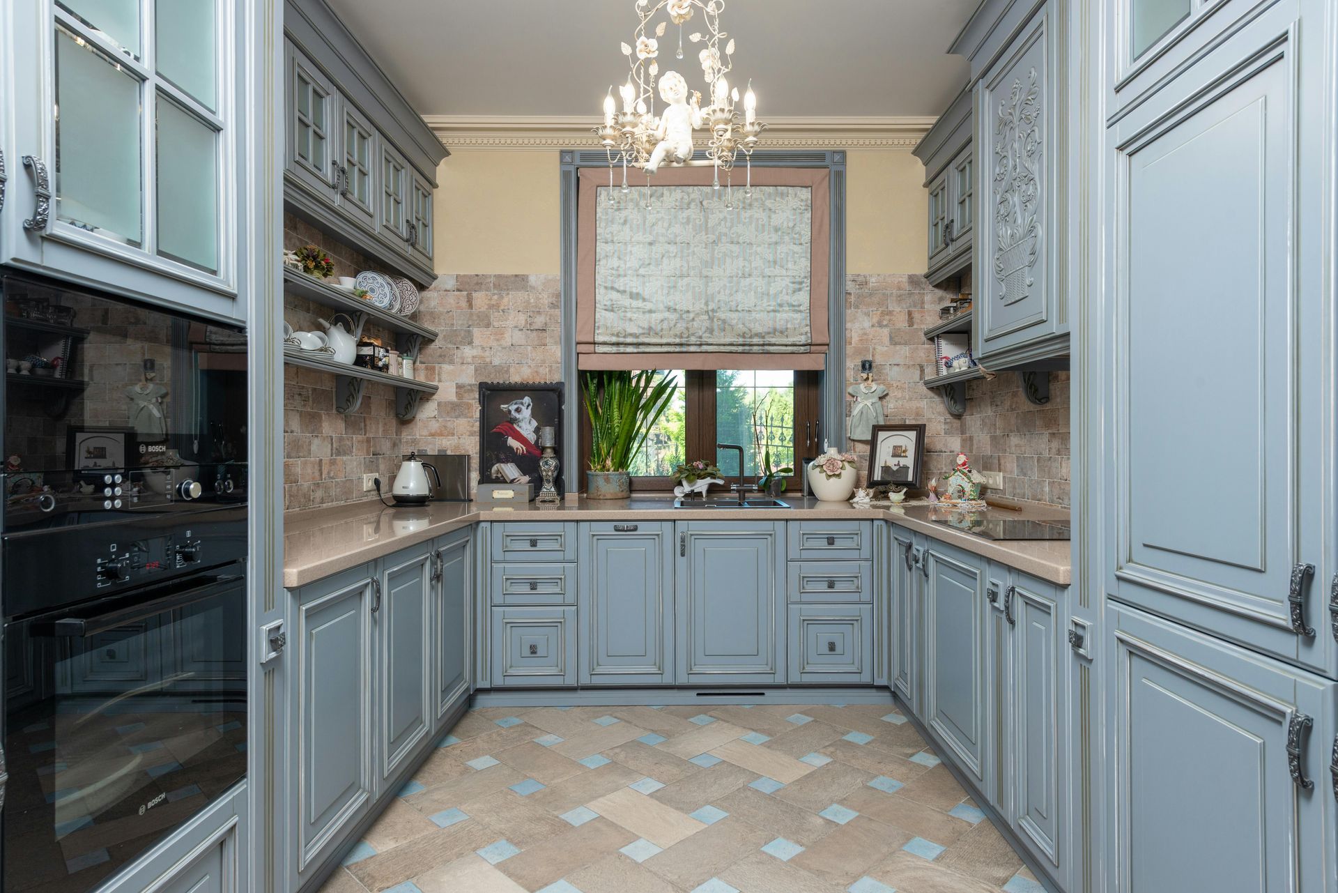 Gray kitchen with cabinets, backsplash, and patterned floor. Window with neutral shade and chandelier.