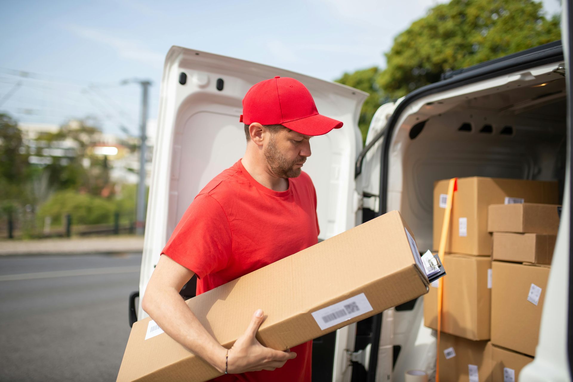 A person in a red uniform and cap holds a rectangular package while standing by the open rear doors of a delivery van.