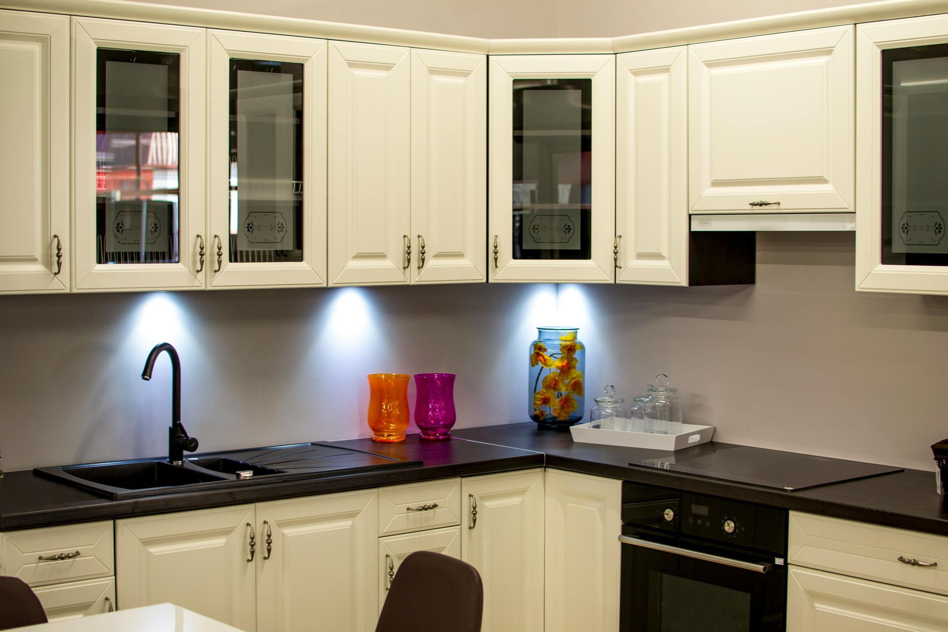 White kitchen cabinets with black countertops and sink. Glass-front cabinets, under-cabinet lighting, and an oven.