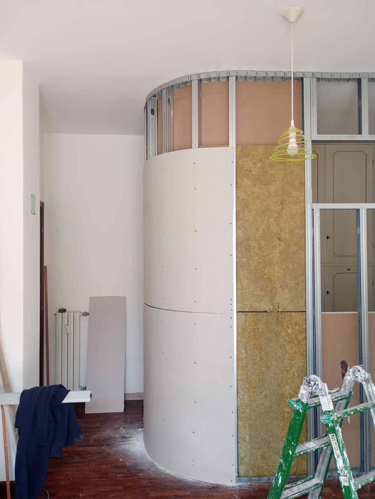 Parete curva in costruzione all'interno di una stanza, con isolamento a vista e scale nelle vicinanze.