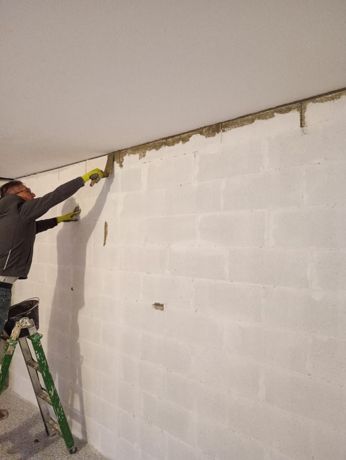 Un operaio su una scala applica materiale isolante o intonaco lungo la parte superiore di un muro di blocchi bianchi.