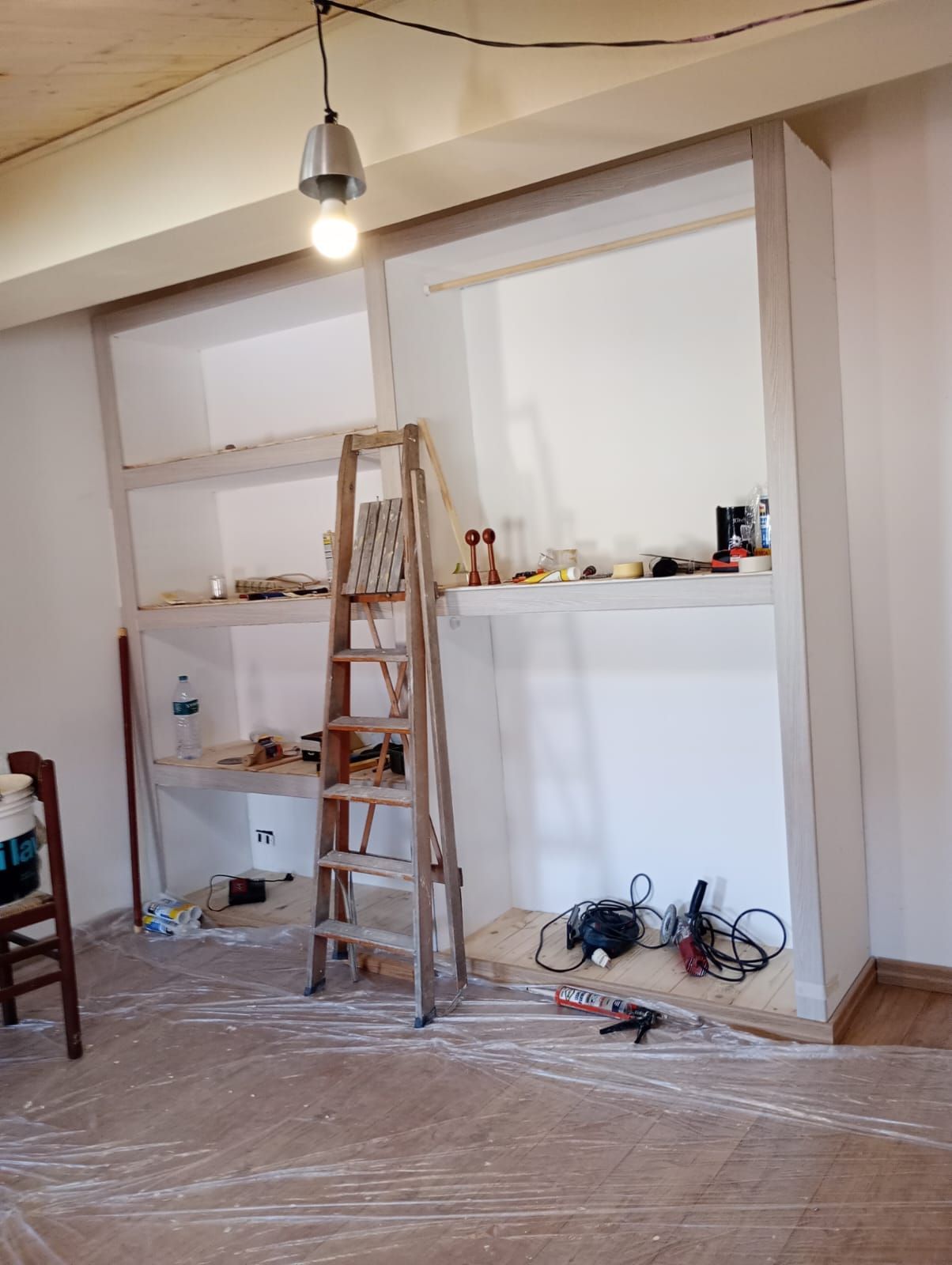 Stanza adibita a ristrutturazione con scala, scaffali, lampadina sospesa e attrezzi su un pavimento ricoperto di plastica.