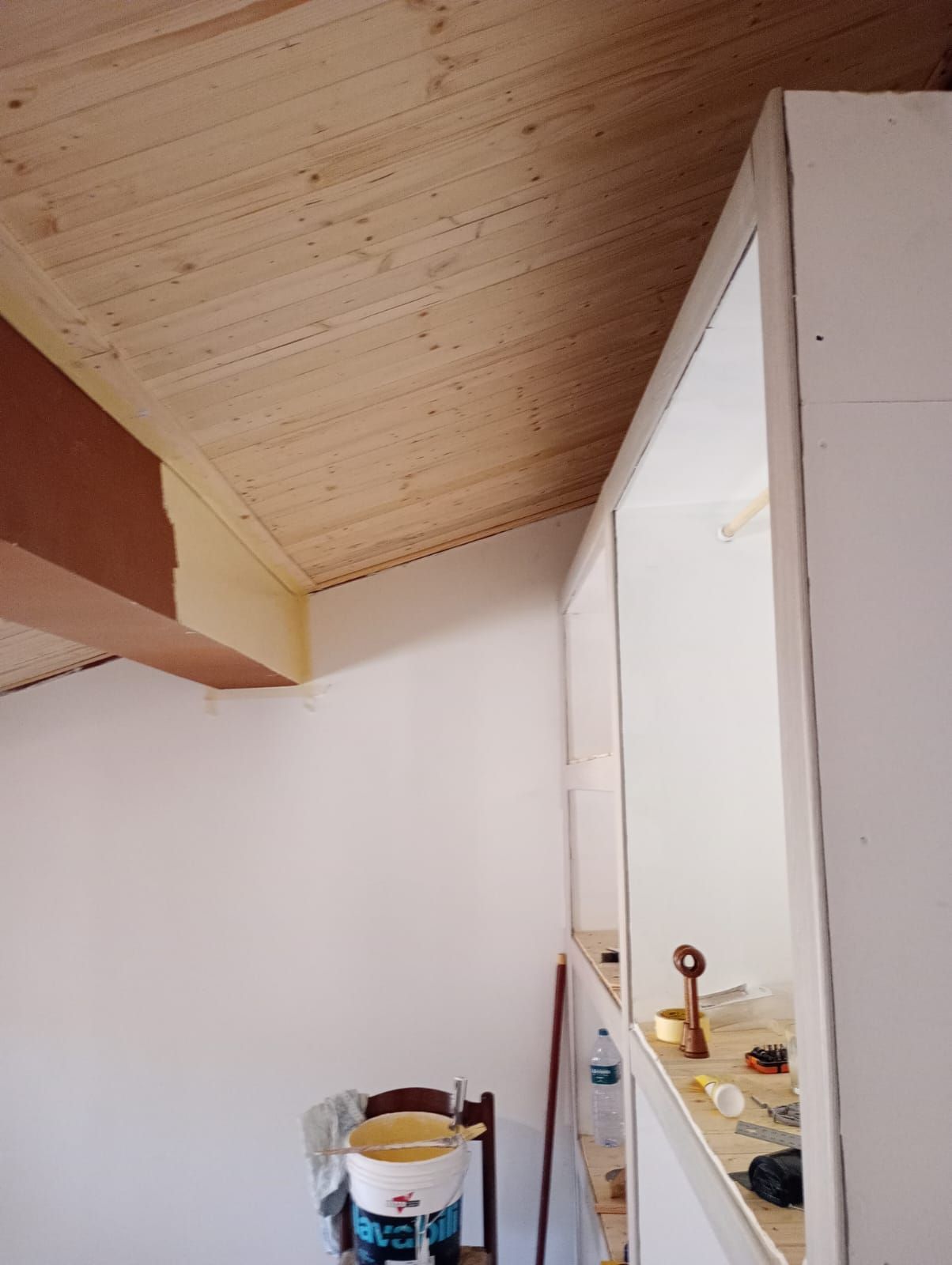Piccola mansarda con soffitto in legno inclinato, pareti bianche e una sedia accanto a una mensola o a un'apertura.