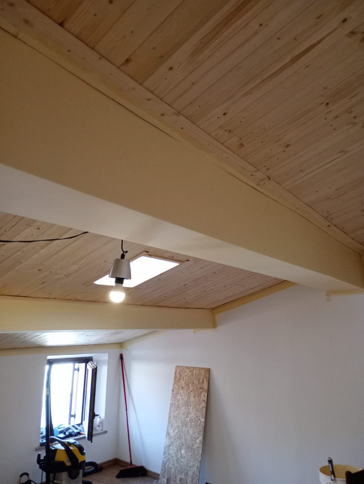 Soffitto a soppalco con pannelli in legno chiaro, travi a vista, lucernario e una stanza sottostante in fase di ristrutturazione.