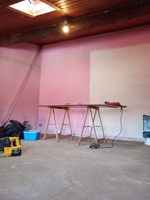 Stanza vuota in fase di ristrutturazione con pareti rosa, pavimento non rifinito, tavoli da lavoro e attrezzi.