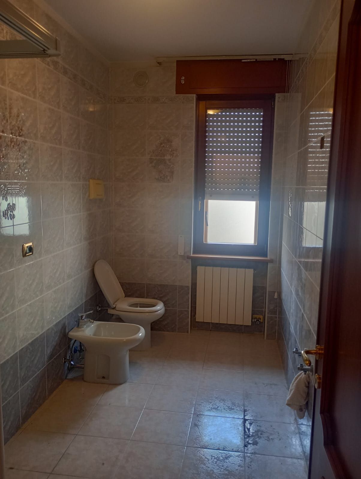 Piccolo bagno piastrellato con WC, bidet, finestra e tenda da doccia; pavimento bagnato visibile.