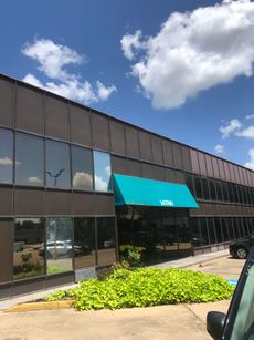 Modern office building with dark glass windows, teal awning, and landscaped entrance under a blue sky.