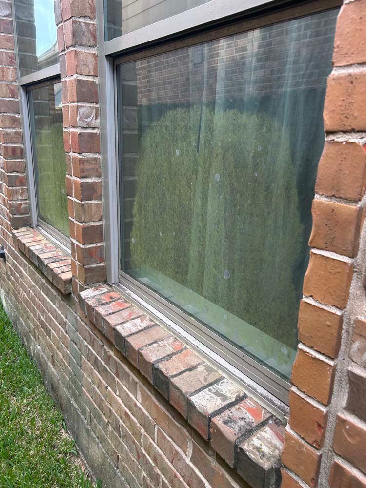 Brick house window with a green curtain and red-brick sill beside a narrow strip of grass