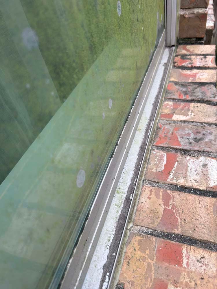 Dirty window track beside a brick walkway, with green-tinted glass and weathered frame.