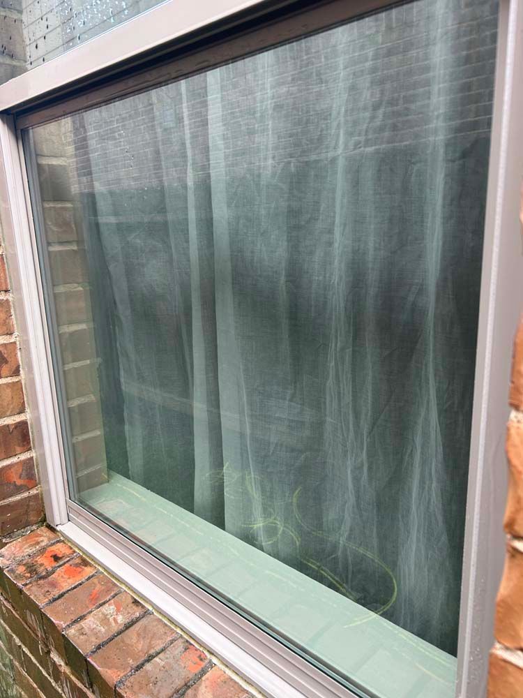 Close-up of a window with a gray curtain, framed by brick and white trim.