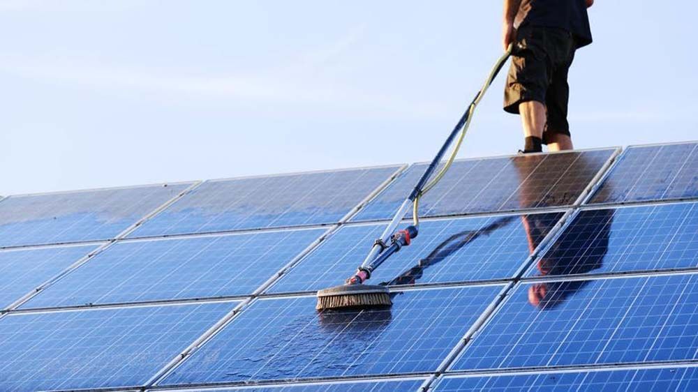 Worker cleaning blue solar panels with a brush on a rooftop, with sky reflected in the panels