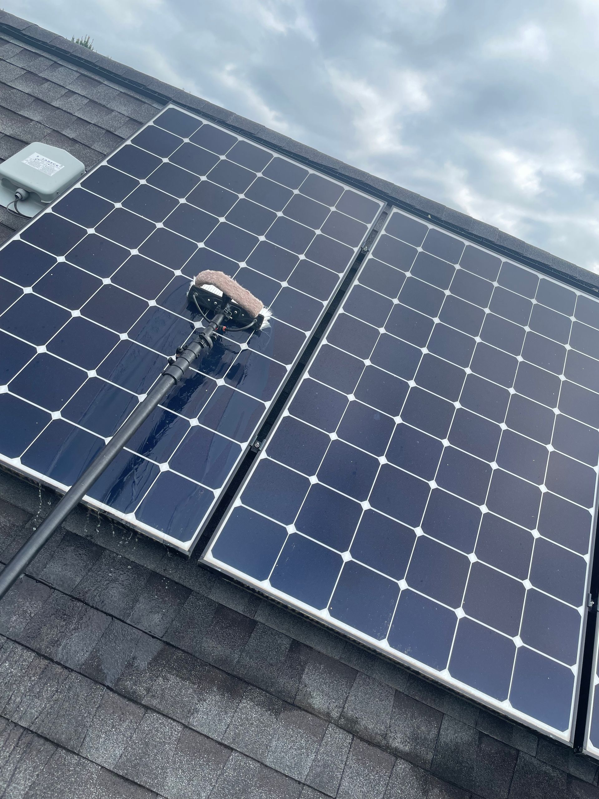 Solar panels on a roof being cleaned with a long pole under a cloudy sky