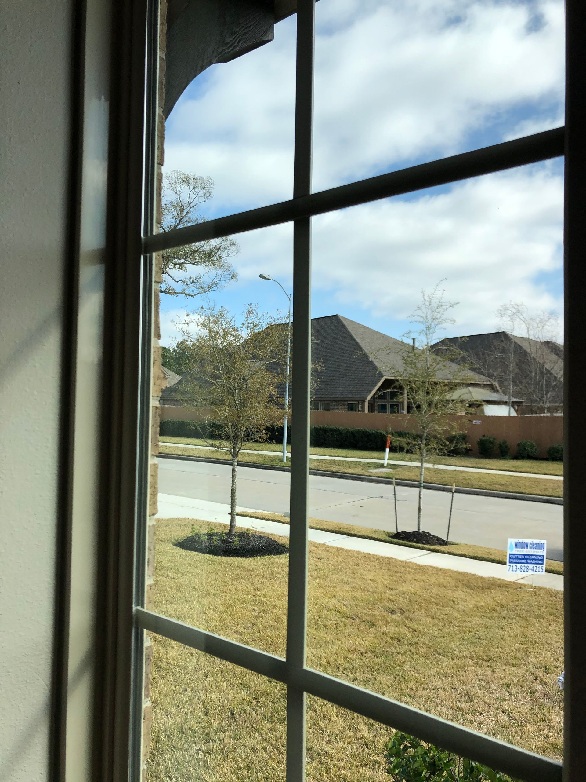 View through a window of a suburban street, sidewalk, lawn, and houses under a partly cloudy sky