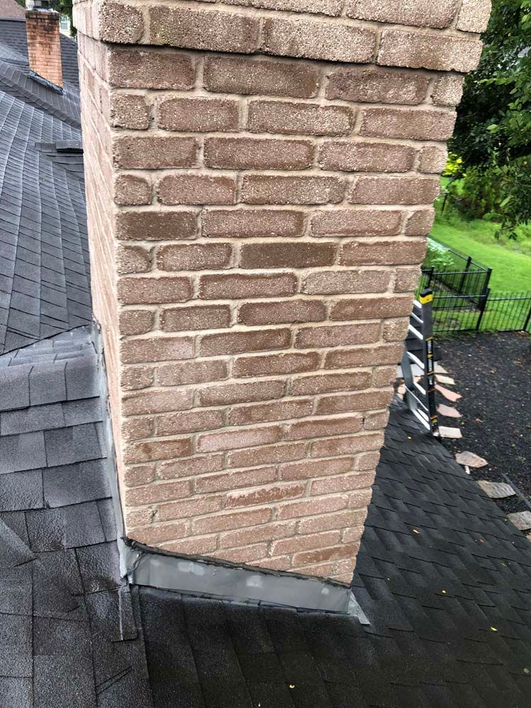 Brick chimney above a dark shingled roof, with a gutter and green trees in the background.